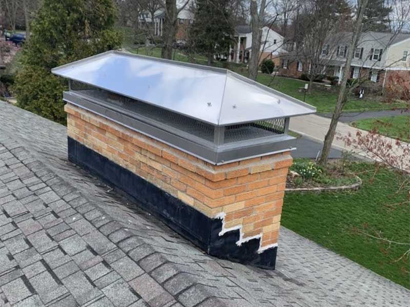 Custom-fitted stainless steel chimney cap on brick chimney - Homes and trees in the background.